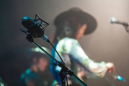 Microphone and musicians at music gig. A closeup view of a microphone and mic stand at a live music event. Blurred performers stand on stage in background. Lead vocalist equipment during gig.の写真素材