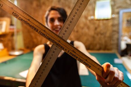 A female clothes maker is standing inside her vintage workshop, holding two wood measuring sticks. Artisan shows tools of trade.の写真素材