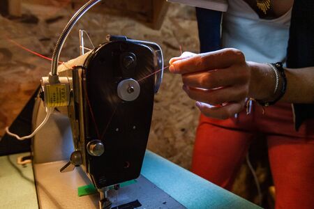 Hands of a young dressmaker are viewed close up, fiddling with red red thread on an industrial...