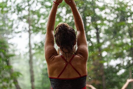 Diverse people enjoy spiritual gathering A young caucasian woman is seen from behind with arms in air, meditating against a blurry forest background with copy space to the sides.の写真素材