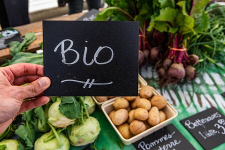 Organic produce at a farmer's market. A first person perspective of a man holding a bio sign over trays of fresh vegetables displayed on a greengrocer's stand during a fair for locally sourced foodの写真素材