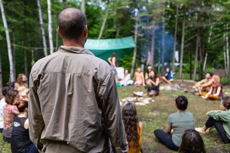 Diverse people enjoy spiritual gathering A middle aged guy with a bald spot is viewed from behind, standing amongst a group of intergenerational people enjoying mystical rituals at a woodland retreatの写真素材