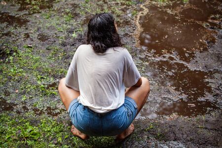 Diverse people enjoy spiritual gathering A young woman wearing a white t shirt and denim jeans is seen crouching barefooted in the mud, contemplating during a mindfulness retreat.の写真素材