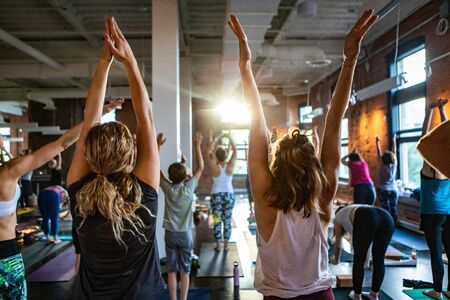 Diverse group of people in yoga class. A view from the back of a gymnasium as participants are seen facing the sun shining through a window during 108 sun salutes (surya namaskar).の写真素材