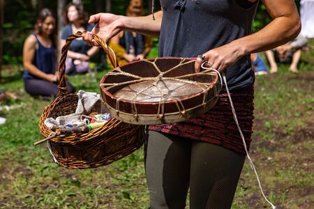 Diverse people enjoy spiritual gathering A close-up view of a young shaman woman holding a native drum and sacred objects in a wicker basket as she walk around a sacred site in a forest clearing.の写真素材