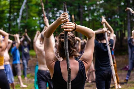 Diverse people enjoy spiritual gathering A close up and rear view of a slim caucasian woman holding a long branch behind her head as multigenerational people practice mindful tai chi in nature.の写真素材