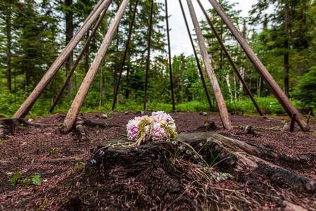 Diverse people enjoy spiritual gathering An offering of flowers is seen closeup, by a sacred tipi (teepee) like structure arranged in a forest clearing during a retreat dedicated to native culture.の写真素材