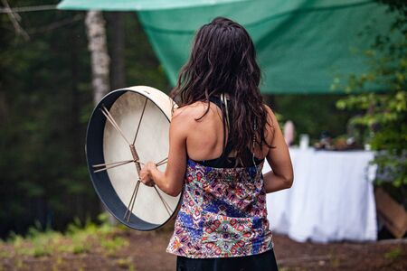 Diverse people enjoy spiritual gathering A caucasian shaman lady is viewed from the rear wearing a colorful native patterned top and playing a sacred drum at a forest campsite.の写真素材