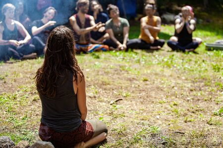 Diverse people enjoy spiritual gathering A close up and rear view of a shaman practitioner sitting on grass in a sacred forest clearing, teaching ancient traditions to blurry people in background.の写真素材