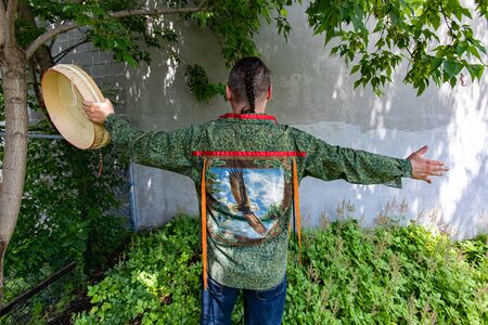 Entertainment at multicultural festival. A close up and rear view of a man wearing a colorful shirt with a sacred eagle feather, symbolizing strength and wisdom, holding a native drum during music gig.の写真素材