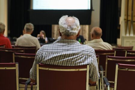 People attend local town hall meeting. A close up and rear view on the back of an elderly man's head, with grey hair and bald spot, during a village council assembly.の写真素材