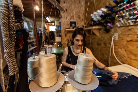A creative clothes designer is viewed sitting at workstation. Young woman uses overlock machine with...
