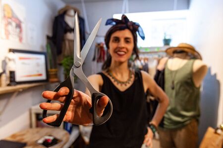 A trendy seamstress is viewed from the front holding a pair of crafting scissors inside an atelier....