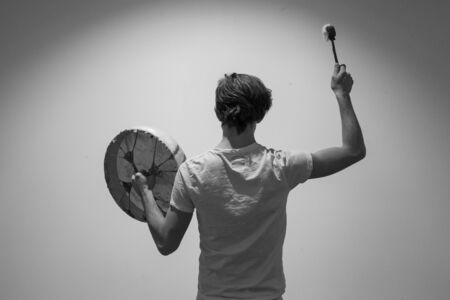 Young man pictured from the back is holding his sacred drum in a hand and raises his drumstick with the other. In the spotlightの写真素材