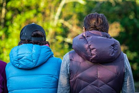Selective focus photo of the behind of two people dressed in warm winter coats and a baseball cap standing in a nature sceneryの写真素材