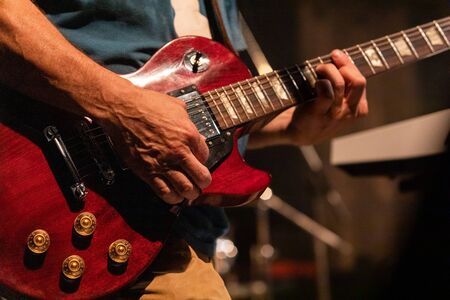 close up and selective focus on mans hands playing his red wooden electric guitar, with a blurry...
