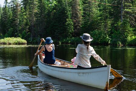 Grandmother with mother and daughter are seen from the back as they riding small wooden boat and Rowing on the lake, blurred trees on the backgroundの写真素材