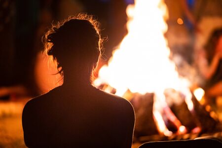 silhouette of a man with long hair sitting in the front of the fire, seen from behind during dark night campfire, blurred backgroundの写真素材