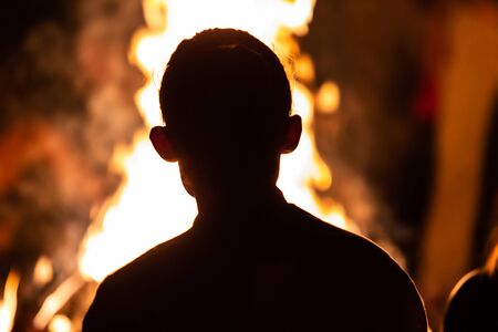 silhouette of an unrecognizable person sitting in the front of the fire, seen from behind during dark night campfire, blurred backgroundの写真素材