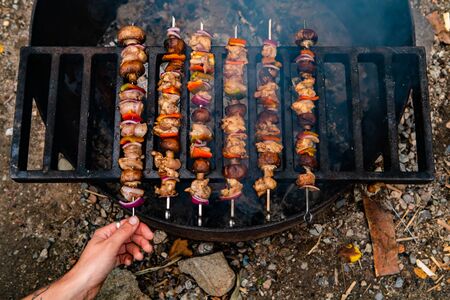 High angle photograph of a large smoking barbecue and a persons hand reaching for slowly grilled skewers with meat and vegetablesの写真素材