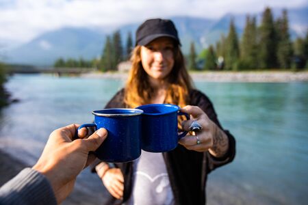 Selective focus of two happy adults spending time together drinking coffee and tea with in the background mountains and a clear riverの写真素材