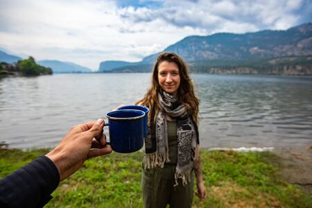 Selective focus of a person clinching a cup of coffee with a young adult female while standing in front of a large lake and mountainsの写真素材