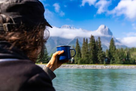 Backside view photograph of adult person standing with cup of coffee or tea while looking at the snowy mountains in the backgroundの写真素材