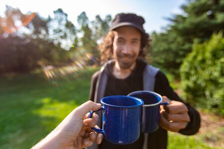 Selective focus of two cheerful people toasting their cups of coffee and tea while standing in a place full of nature scenery and surroundingsの写真素材