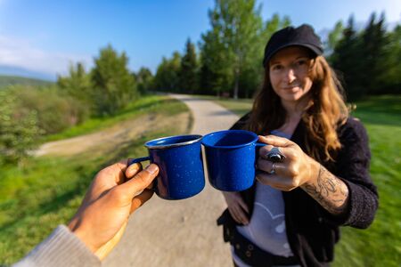 Selective focus of two adults clinching their cups of warm coffee and tea while spending time together in a nature scenery on a sunny dayの写真素材