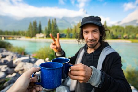 Selective focus of young middle aged couple drinking coffee and tea while standing in front of a blue river and mountains in the backgroundの写真素材