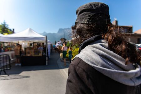 A man wearing a black jacket and baseball cap is seen from the back at a street fair during summer, blurry stalls are in background with copy spaceの写真素材