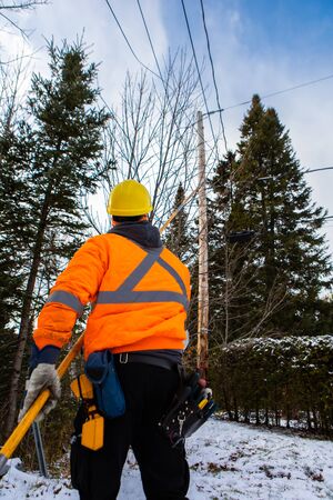 An optical fiber engineer is seen from the back, working under a telegraph pole outdoors in winter,...
