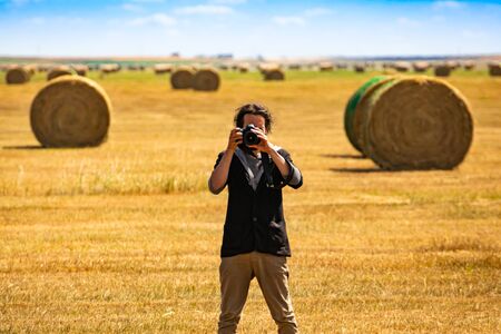 Wide angle view of male photographer standing in an agricultural field with bales of golden hay, shooting at camera with a DSLR camera. の写真素材
