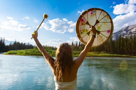 A shamanic woman is seen celebrating nature and life from the back, holding a sacred drum and beater in the air against a peaceful natural landscapeの写真素材
