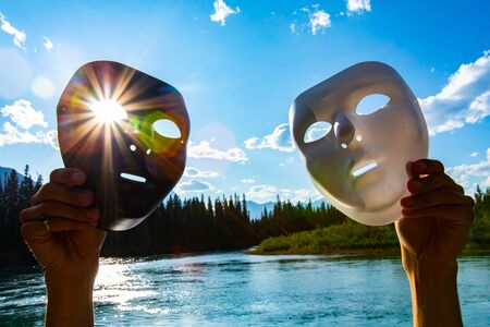 Light and dark concept in nature as a person holds two masks against the sun over a large lake by mountains, with lens flare as rays shine through eyeの写真素材