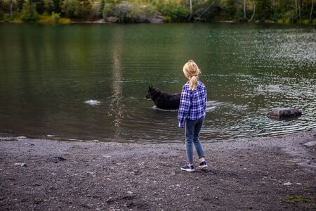 little girl in a checkered shirt watching her cute dog play in the green and transparent water of a...