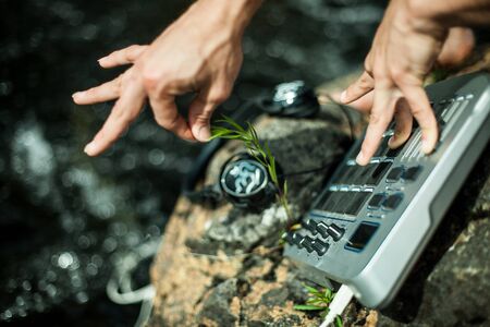 Caucasian male performing music using sampler in the nature sitting on a rock near the banks of crystal clear river. Sunny relaxing scenery.の写真素材
