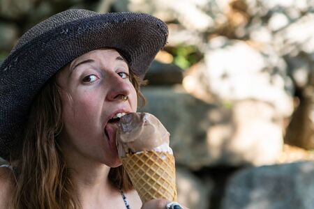 A close up and front portrait of a caucasian with pierced nose licking an ice cream cone. sitting in the shade near some blurry rocksの写真素材