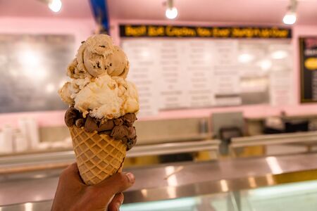 A close up and first person perspective of a man holding a waffle cone with scoops of ice cream at shop, with blurry menu in the backgroundの写真素材