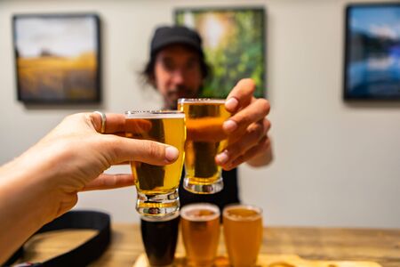 two beer glasses cheering close up selective focus point of view, man in the bar background, flight of Craft of different Beers glasses on Wooden Trayの写真素材