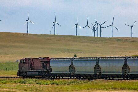 Row of metallic wagons of a Canadian freight train running between green fields in the countryside. Wind turbines in the background. Red locomotive.の写真素材