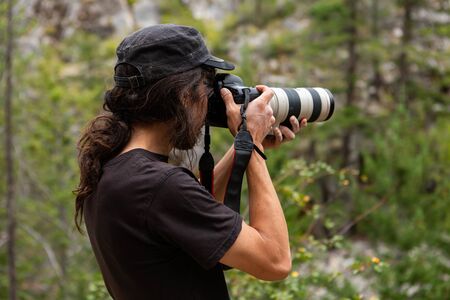 A close up and side shot of a wildlife photographer at work, wearing a black t shirt and cap, using...