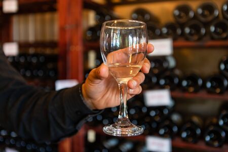 A glass of white wine on a man hand selective focus close up view against wine bottles on storage...