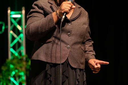 Midsection of woman standing and holding microphone while pointing her finger and giving speech on stage in auditorium hallの写真素材