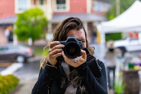 Portrait of professional female photographer wearing warm clothing and taking photograph from her camera on city street in summer seasonの写真素材