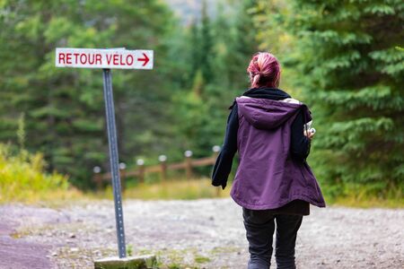 Rear view of woman with dyed hair wearing warm clothing and walking alone by french bike return text direction board against trees in forestの写真素材