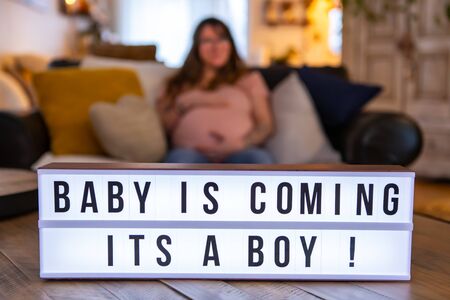 A selective focus shot on a cinematic LED lightbox with the message baby is coming its a boy, a blurry pregnant woman is seen sitting in backgroundの写真素材
