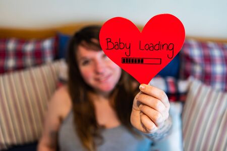 A selective focus shot of a pregnant woman holding a red heart with message baby loading as she prepares for valentine childbirth, with copy spaceの写真素材