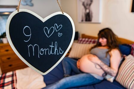 A selective focus shot of a heart chalkboard sign saying 9 months. with a blurry pregnant woman...