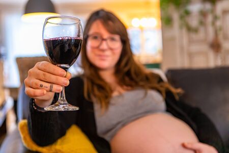 A selective focus view of a pregnant woman in third trimester enjoying a glass of red wine at home, risking health of unborn baby, copy space to rightの写真素材